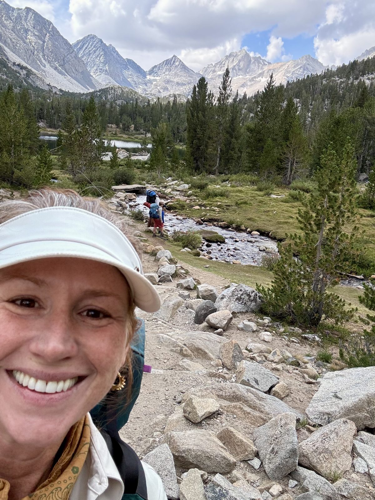Sonya smiling in a white sun visor in a selfie, with her family hiking beside a stream in an alpine valley behind her