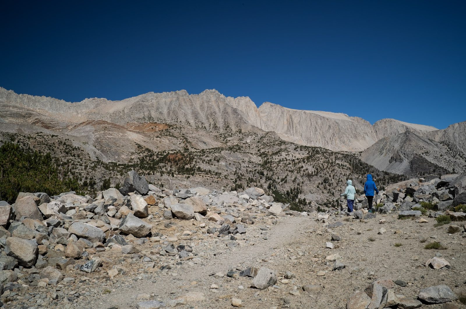 Two boys in rain jackets hiking up a rocky trail toward a high mountain pass under a deep blue sky