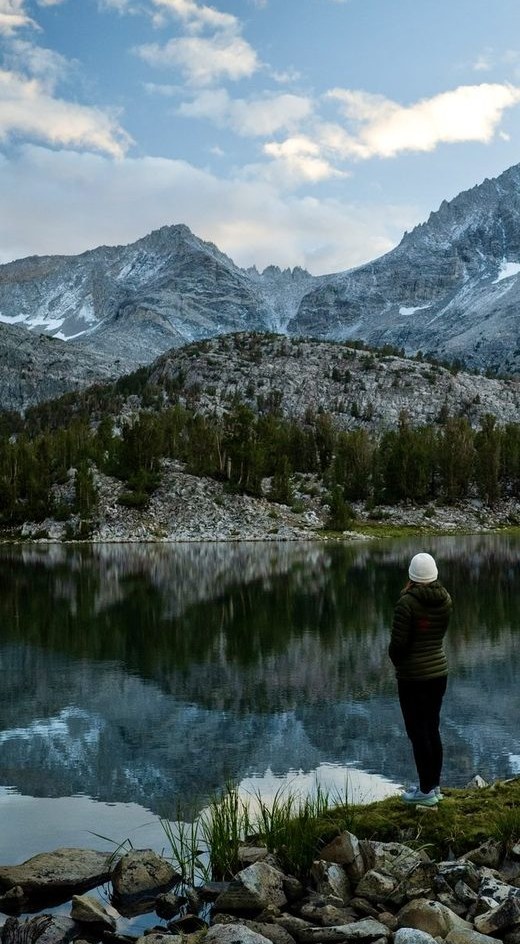 A person in a white beanie standing at the edge of a still alpine lake, mountains reflected in the water