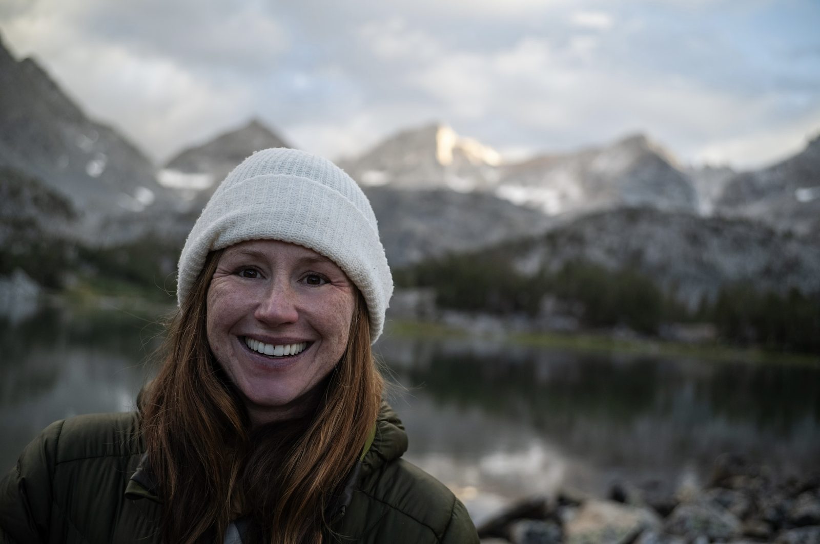 Sonya smiling in a white beanie and puffy jacket beside an alpine lake at dusk