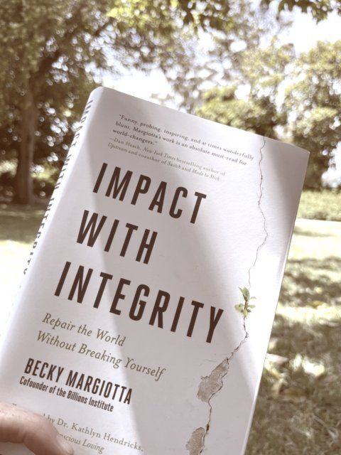 Sonya holding her copy of Impact with Integrity by Becky Margiotta, outdoors under trees